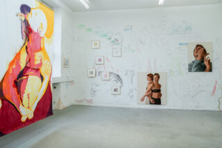 Ein Kunstatelier mit einem großen bunten Gemälde auf Stoff, gerahmten Skizzen an der Wand und zwei Fotoporträts mit Bleistiftzeichnungen auf dem Boden, die an einer weißen Wand lehnen.