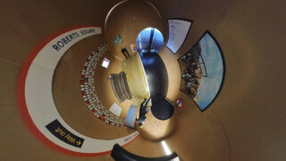 Eine verzerrte Panoramaansicht eines Innenraums mit einem Schild für „Roberts Square“, Bänken, einer gefliesten Wand und einem großen ausgestellten Foto.