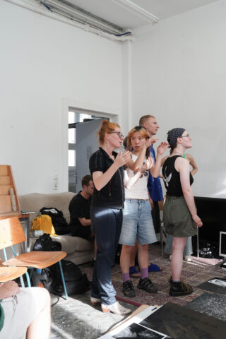 Eine Gruppe von Menschen steht und gestikuliert, während andere in einem hellen Raum mit weißen Wänden, Möbeln und verschiedenen verstreuten Gegenständen sitzen.