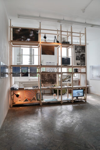 Ein großes Holzregal voller gerahmter Fotos, kleiner Gegenstände und Dekorationen steht vor einer weißen Wand in einem Galerieraum mit Betonböden und großen Fenstern.