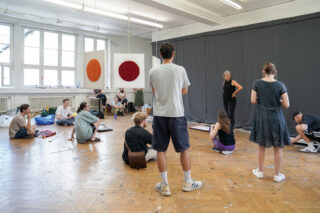 Eine Gruppe von Menschen versammelt sich in einem Kunstatelier. Einige sitzen auf dem Boden, andere stehen und hören einer Frau zu, die vor einem dunklen Vorhang spricht. Künstlerutensilien und Wandbehänge sind zu sehen.