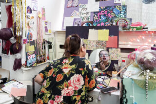 Eine Person mit langen Haaren und einem geblümten Oberteil trägt Lippenstift vor einem überladenen Kosmetikspiegel auf, der von bunten Notizen, Make-up und Dekorationen umgeben ist.