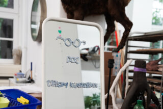 Ein Spiegel mit weißem Rahmen und Text und Illustrationen auf der Oberfläche, platziert auf einem unordentlichen Tisch in einem Kunstatelier, mit sichtbaren verschiedenen Utensilien und Geräten.