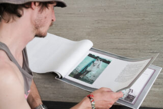 Eine Person mit Hut und Tanktop sitzt auf einer Betonfläche und liest eine große aufgeschlagene Zeitschrift oder ein Buch mit einem Foto und Text.