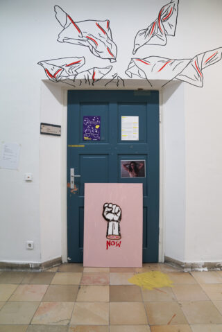 Eine blaue Doppeltür mit Plakaten, ein rosa Protestschild mit erhobener Faust und dem Wort „JETZT“ sowie schwarz-rote Zeichnungen von Blättern oder Blütenblättern über der Tür.