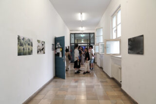 Eine Gruppe von Menschen steht und unterhält sich in einem Flur mit weißen Wänden, gefliesten Böden, offenen Fenstern und an den Wänden hängenden Fotos.