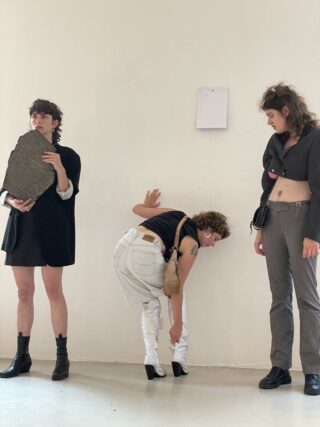 Drei Menschen stehen vor einer weißen Wand. Einer hält eine große Platte, ein anderer beugt sich hinunter, um seinen Schuh zu richten, und der dritte steht mit ausgestreckten Händen da und sieht die anderen an.