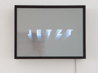 Auf einem schwarz umrahmten Bildschirm wird das Wort „JETZT“ in weißen Buchstaben mit einem nachlaufenden Unschärfeeffekt auf grauem Hintergrund angezeigt.