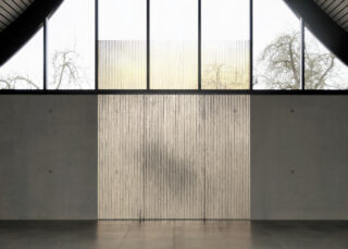 Große Fenster mit vertikalen gerippten Glasplatten lassen diffuses Licht in einen minimalistischen Innenraum mit Betonwänden und Fliesenboden. Draußen sind kahle Bäume sichtbar.