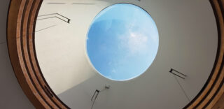 Blick nach oben durch ein rundes Oberlicht mit in das Glas eingraviertem Text: „OBJEKTE IM WELTRAUM SIND GRÖSSER ENTFERNT, ALS SIE ERSCHEINEN.“ Durch das Fenster ist blauer Himmel sichtbar.