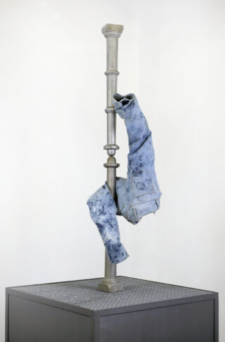 Eine Bluejeans, die wie Beine geformt ist, hängt kopfüber von einer Metallstange, die auf einer quadratischen Plattform montiert ist.