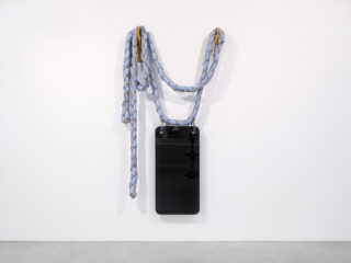 Ein geflochtenes Stoffseil in Blau- und Grautönen ist über ein schwarzes rechteckiges Paneel drapiert, das an einer weißen Wand angebracht ist.