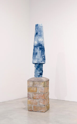 Eine Skulptur eines mit blauem, batiktem Stoff bedeckten Objekts, das aufrecht auf einem rechteckigen Ziegelsockel in einer Galerieecke mit weißen Wänden steht.