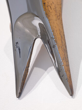 Nahaufnahme eines glänzenden, dunklen, V-förmigen skulpturalen Objekts mit glatter Oberfläche und rostigen, strukturierten Kanten, das an einer weißen Wand montiert ist.