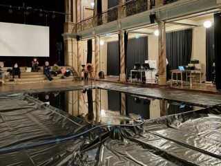 Eine Gruppe von Menschen sitzt auf einer Bühne in der Nähe eines großen Wasserbeckens, das mit schwarzen Plastikplanen abgedeckt ist, in einem hohen, reich verzierten Raum, in dem Computer und Geräte sichtbar sind.