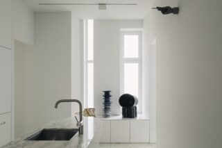 Minimalistische Küche mit Marmorarbeitsplatte, Edelstahlspüle und dekorativen Vasen auf einem weißen Sims neben hohen Fenstern; hoch oben an der Wand hängt die Skulptur einer zeigenden Hand.