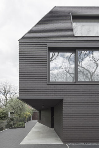 Modernes Gebäudeäußeres mit dunkelgrauer Schindelverkleidung, großen Fenstern, in denen sich Bäume spiegeln, und einem Überhang über einem Gehweg.