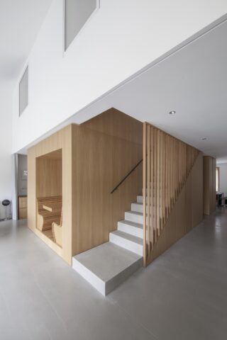 Minimalistisches Interieur mit einer Holztreppe mit vertikalen Lamellen, hellgrauen Betonstufen und einer eingebauten Holznische mit Sitzgelegenheiten unter der Treppe.