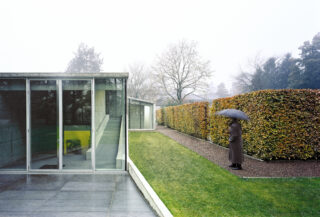 Eine Person mit einem Regenschirm geht an einem nebligen Tag einen Weg neben gepflegten Hecken in der Nähe eines modernen Glasgebäudes entlang.