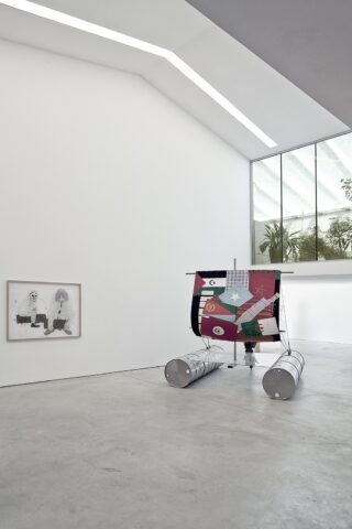 Ein moderner Galerieraum mit einem gerahmten Kunstwerk an der Wand und einer farbenfrohen, geometrischen Skulptur auf dem Boden, die an ein kleines Boot mit Metallpontons erinnert.