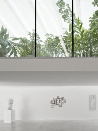 Eine minimalistische Kunstgalerie mit weißen Wänden zeigt eine Büste auf einem Sockel und gerahmte Kunstwerke; große Fenster darüber geben den Blick auf üppiges grünes Laub draußen frei.