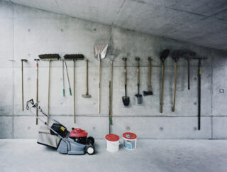 Eine Reihe Gartengeräte hängt an einer Betonwand über einem Rasenmäher von Honda und zwei Farbeimern auf einem nackten Boden.