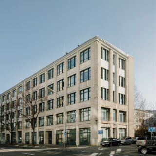 Ein vierstöckiges beiges Bürogebäude mit großen Fenstern auf jeder Etage, gelegen an einer Straßenecke mit parkenden Autos und kahlen Bäumen in der Nähe, unter einem klaren blauen Himmel.