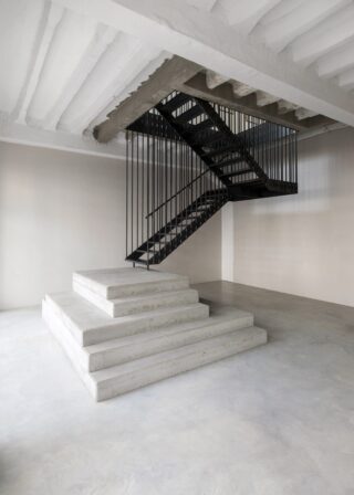 Eine moderne Treppe im Industriestil mit schwarzem Geländer führt von einer zentralen, vierstufigen Betonplattform in einen minimalistischen, offenen Innenraum mit freiliegenden Balken hinauf.