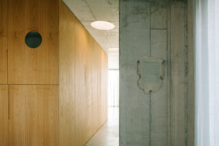 Ein minimalistisches Interieur mit einer holzgetäfelten Wand auf der linken Seite und einer freiliegenden Betonsäule mit einer abgeplatzten Stelle auf der rechten Seite, beleuchtet durch natürliches Licht.