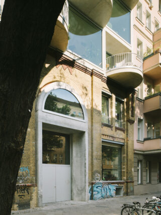 Ein modernes Wohnhaus mit großen Glasfenstern, Balkonen, gelber Backsteinfassade und Graffiti an den unteren Wänden; davor sind Fahrräder abgestellt.