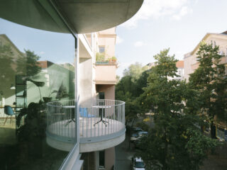 Von einem runden Wohnungsbalkon mit einem Tisch und zwei Stühlen hat man Ausblick auf eine von Bäumen gesäumte Straße; in großen Glasfenstern sind Spiegelbilder des Balkons und des Innenraums sichtbar.
