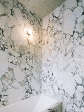 Badezimmerecke mit großen weißen und grauen Marmorfliesen, einer eingebauten Badewanne und einer runden Wandleuchte darüber.