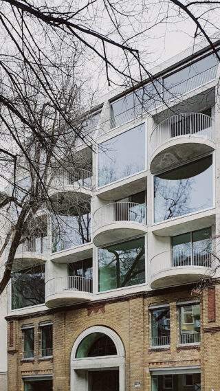 Modernes Mehrfamilienhaus mit großen Glasfenstern und abgerundeten Balkonen, errichtet über einer älteren Backsteinfassade. Im Vordergrund sind blattlose Äste zu sehen.