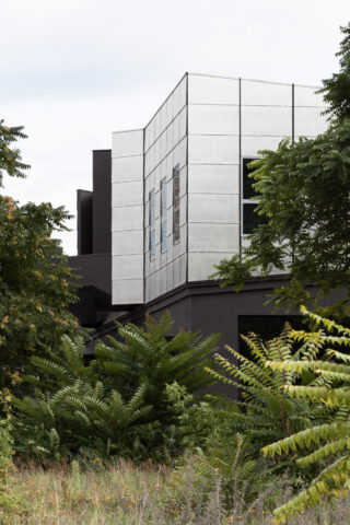 Ein modernes Gebäude mit einer metallischen oberen Fassade und dunklen unteren Wänden, die teilweise durch dichtes grünes Laub im Vordergrund verdeckt werden.