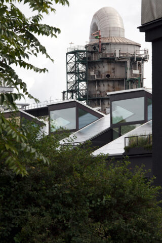 Hinter modernen, eckigen Gebäuden und grünem Laub steht eine große Industriestruktur mit Metallgerüst und geschwungener Spitze.