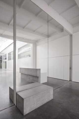 Minimalistisches Interieur mit Betonbänken, großen Spiegeln und weißen Wänden unter einer hohen Decke mit freiliegenden Balken.