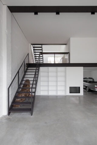 Modernes, minimalistisches Interieur mit Metalltreppe, weißen Wänden, eingebauten Regalen, einem Kamin und einem teilweise sichtbaren Oldtimer in einem angrenzenden Garagenbereich.