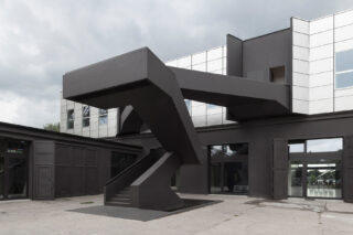 Ein modernes Gebäude mit einer markanten, eckigen schwarzen Außentreppe und spiegelnden Fenstern unter einem bewölkten Himmel.