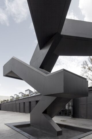 Eine moderne Außentreppe mit eckigem, geometrischem Design aus dunklem Material steht in einem offenen Bereich mit Gebäuden und einem bewölkten Himmel im Hintergrund.