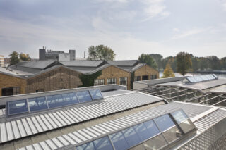 Industriegebäude mit Backstein- und Metallfassaden und großen Oberlichtern auf den Dächern, umgeben von Bäumen unter einem teilweise bewölkten Himmel.