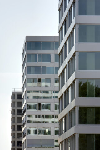 Drei moderne Hochhäuser mit Glas- und Metallfassaden stehen nebeneinander unter klarem Himmel. In den Fenstern sind Spiegelungen der umliegenden Bäume zu sehen.