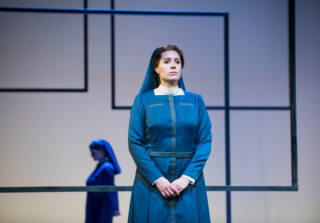 Zwei Frauen in blauen Uniformen stehen auf einer minimalistischen Bühne mit geometrischen Rahmen; eine blickt nach vorne, die andere geht seitwärts im Hintergrund.