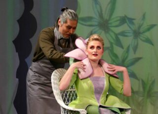 Ein Mann steht hinter einer sitzenden Frau in einem Blumenkostüm und richtet ihren Kragen vor einem floralen Hintergrund.