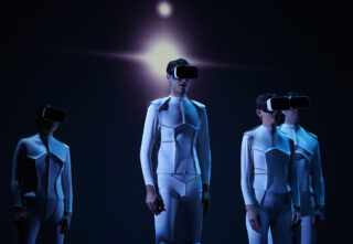 Vier Personen in weißen futuristischen Ganzkörperanzügen und VR-Headsets stehen vor einem dunklen Hintergrund mit einem hellen Licht über ihnen.