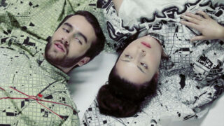 Ein Mann und eine Frau liegen nebeneinander auf einer weißen Fläche. Beide tragen gemusterte Kleidung mit geometrischen und kartenähnlichen Mustern und blicken in unterschiedliche Richtungen.