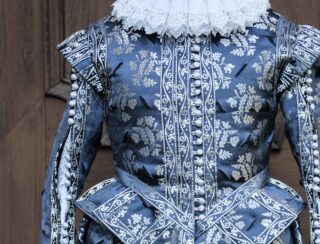 Nahaufnahme eines kunstvollen historischen Kleidungsstücks in Blau und Silber mit Blumenmuster, Knöpfen auf der Vorderseite, strukturierten Schultern und einem weißen Spitzenkragen.