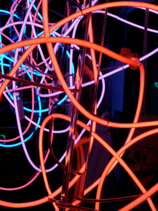 Helle Neonröhren in den Farben Orange, Pink und Blau sind vor einem dunklen Hintergrund verwickelt und ineinander verschlungen.