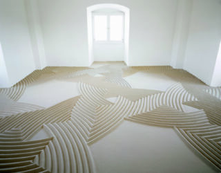 Ein minimalistischer weißer Raum mit einem Fenster; der Boden ist mit Sand bedeckt, der in präzisen, geometrischen, wellenartigen Mustern angeordnet ist.