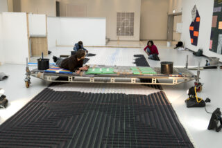 Drei Personen arbeiten in einem geräumigen, hellen Raum an einer großen Bodeninstallation mit Gittermustern; Materialien und Werkzeuge sind um sie herum verteilt.