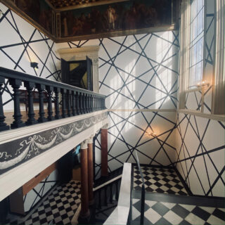 Innenansicht eines historischen Gebäudes mit schwarz-weiß kariertem Boden, geometrischen Wandmustern, großen Fenstern und einem oberen Balkon mit Balustrade.